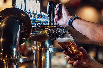 A bartender pours amber draft beer from a shiny tap into a glass, with foam forming at the top. The scene captures a close-up of the pouring process in a bar setting