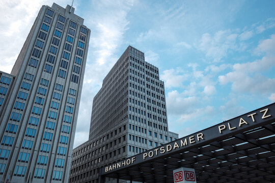 Low-angle outdoor view of modern high-rise office towers and the entrance structure of Potsdamer Platz station under a partly cloudy blue sky in Berlin, Germany.  
