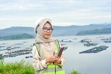 A female fish farm manager wears a safety vest and carries a book to check the work.