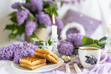 a cup with a hot drink. Breakfast with waffles and cakes, a bouquet of lilacs in a wicker basket