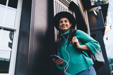 Smiling brunette woman, 30, listens to music and holds smartphone while walking, representing joy,...
