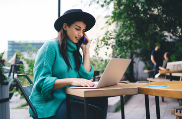 Focused woman, 30, talking on smartphone while typing on laptop outdoors, multitasking during...