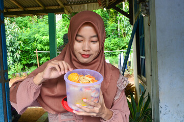 Beautiful Asian woman smiling in pink dress and hijab showing gesture while eating Eid snacks, isolated on white background