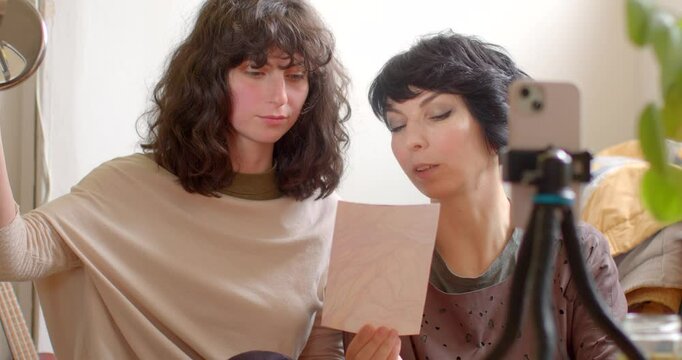 In a cozy indoor setting, two women have an intimate discussion about artwork and makeup techniques, using a mirror and a piece of art for inspiration in their creative exchange