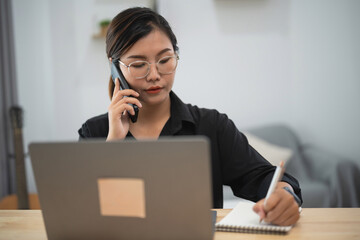 Professional woman engaged in phone conversation while taking notes at a stylish desk with laptop...