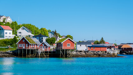 Naklejka premium Colorful wooden cabins line the tranquil waters of Lofoten Norway, embodying the scenic charm of coastal living. Surrounded by lush greenery, this picturesque spot invites relaxation and exploration.