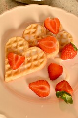 Delicious heart-shaped waffles topped with fresh strawberries and condensed milk. Romantic homemade breakfast served on a plate.