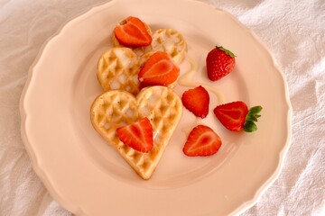 Delicious heart-shaped waffles topped with fresh strawberries and condensed milk. Romantic homemade breakfast served on a plate.
