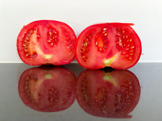 Two fresh red tomatoes are cut in half, revealing their juicy interior filled with seeds. This display highlights their vibrant color and glossy texture against a reflective background.