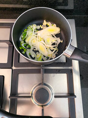 Fresh bell peppers, leeks, and cherry tomatoes are being saut&eacute;ed in a black pan on a stove. Steam rises, indicating a delicious meal is in progress in a bright kitchen.