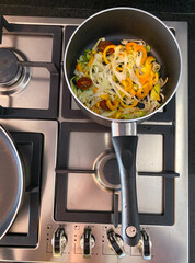 Fresh bell peppers, leeks, and cherry tomatoes are being saut&eacute;ed in a black pan on a stove. Steam rises, indicating a delicious meal is in progress in a bright kitchen.