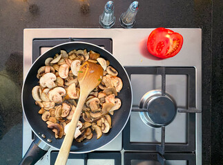 Fresh mushrooms are sliced and saut&eacute;ed in a skillet on a sleek stovetop. A wooden spoon is used to stir the mushrooms as they cook, highlighting the culinary process.