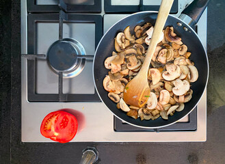 Fresh mushrooms are sliced and saut&eacute;ed in a skillet on a sleek stovetop. A wooden spoon is used to stir the mushrooms as they cook, highlighting the culinary process.
