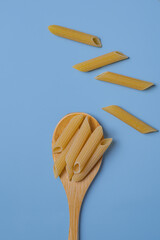 Raw penne pasta on blue background. Uncooked penne on a wooden spoon with scattered pasta over blue. Minimal flat lay composition.