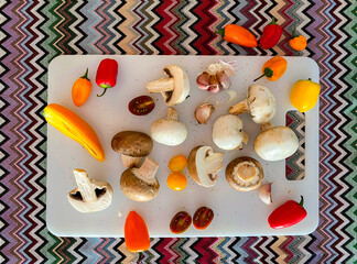 Freshly harvested mushrooms are surrounded by an array of colorful peppers, tomatoes, and garlic, all resting on a vivid patterned fabric, creating a lively kitchen atmosphere.