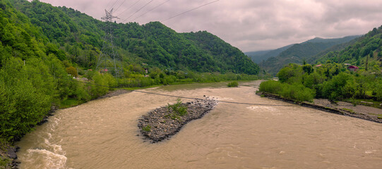 Green hills frame the winding river in Batumi, Adjara, Georgia. Gentle clouds hover over the landscape as nature thrives in this serene setting. The atmosphere is tranquil and inviting.