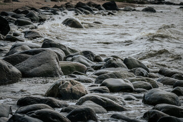 Ocean wave splashes over a dark coastal rock, capturing motion and texture in the clear water under cloudy skies.
