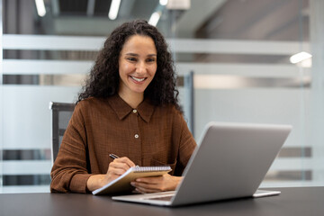 Fototapeta na wymiar A woman with curly hair smiles while writing notes during an online meeting in the office. Her laptop sits open in front of her.