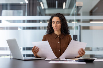 A businesswoman in a brown shirt intently reviews documents at her desk. A laptop, calculator, and...