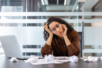 Obraz premium A frustrated businesswoman sits at her desk surrounded by crumpled paper, appearing overwhelmed. She looks tired after a long day.