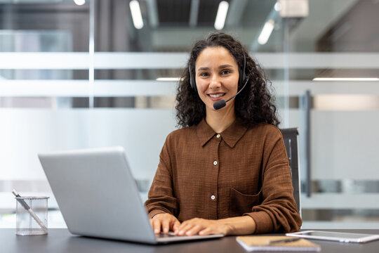 A smiling customer service representative wearing a headset in a modern office setting, using a laptop at a desk.