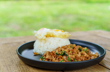 Stir-fried pork and basil leaves with rice and a fried egg served on black plate on wooden table with green background in restaurant. Popular Thai  street food.