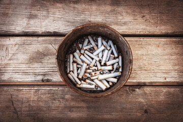 A metal container filled with numerous cigarette butts and ash sits on a weathered wooden surface, viewed from directly above. It's a stark photo of an unhealthy habit's aftermath.