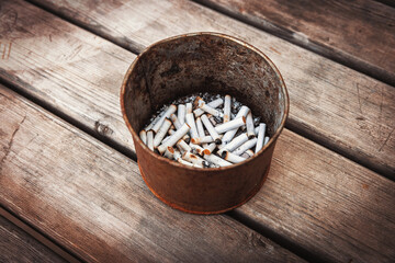 A metal container filled with numerous cigarette butts and ash sits on a weathered wooden surface. It's a stark photo of an unhealthy habit's aftermath.