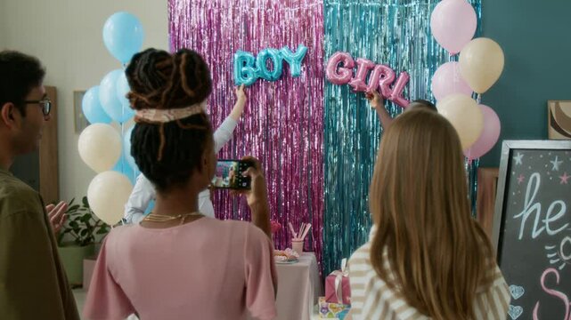 Medium full shot of happy multiethnic expecting couple pointing to blue and pink boy or girl balloons at reveal party, while friends are guessing baby gender and filming memorable moment on smartphone