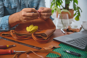 Shoemaker sewing a leather bag in his workshop using needle and thread, surrounded by tools and...