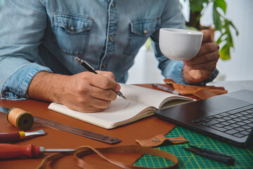 Leather craftsman drinking coffee and writing notes on notebook in his workshop