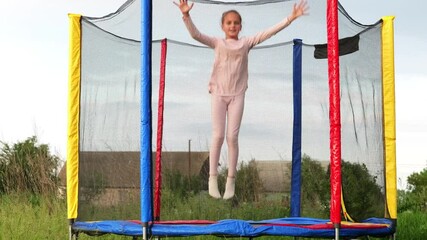 A smiling young girl in pink clothing jumps high on a trampoline with a safety net in a rural outdoor setting.