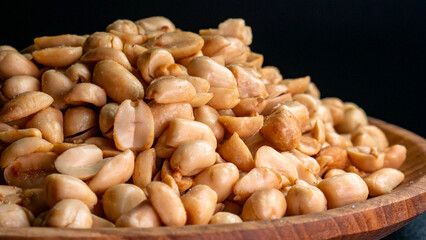 Detail of crispy and salty shelled roasted peanuts in a wooden plate with black background, suitable for snacks, Eid al-Fitr, or culinary concept themes.