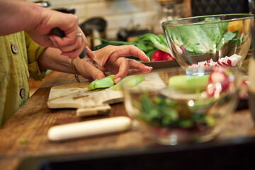 Woman slicing a cucumber on a wooden cutting board in the kitchen, close-up of hands