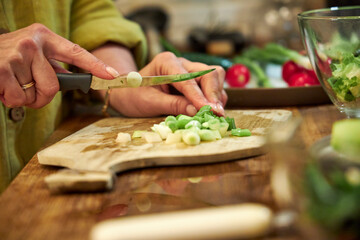 Woman chopping green onions on a wooden cutting board in the kitchen, close-up of hands
