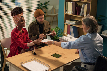 Young adult Black woman and young adult Caucasian man sitting together receiving documents from middle aged Caucasian female social worker in modern office setting