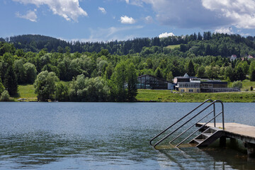 Scenic Lakeside View with Forest and Buildings. A serene lakeside scene with a forested hillside...