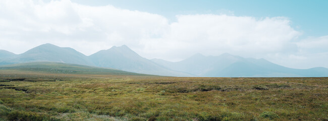 A distant view of the Strickeen mountain range stands beyond the sprawling green landscape of County Kerry, Ireland, under a soft, cloudy sky.