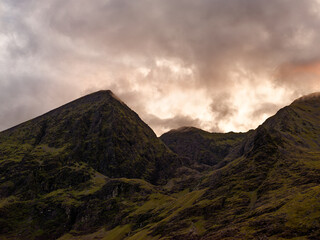Carrauntoohil, the highest peak in Ireland, towers against a backdrop of swirling clouds as dusk approaches. The vibrant landscape displays lush green hills and rocky outcrops.