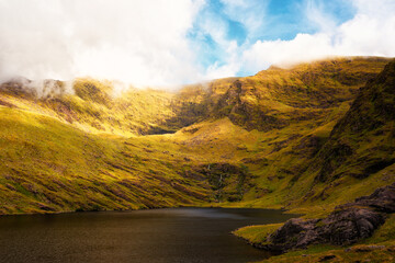Scenic daytime showcases Lough Callee, nestled within green mountains in County Kerry, Ireland. Sunlight illuminates the mountainsides, with light clouds partially obscuring the peaks.