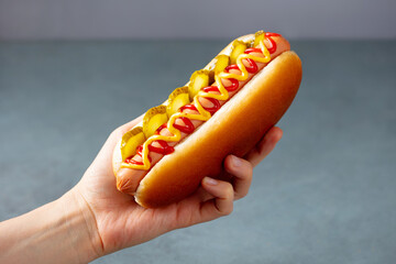 A woman's hand holds a hot dog with ketchup, mustard, pickled cucumbers. Grey background. Close up.