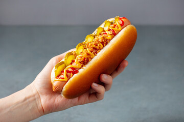 A woman's hand holds a hot dog with ketchup, mustard, pickled cucumbers and fried onions. Grey background. Close up.