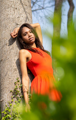 Mujer joven morena de Colombia, con vestido rojo apoyada en un tronco con bokeh verde