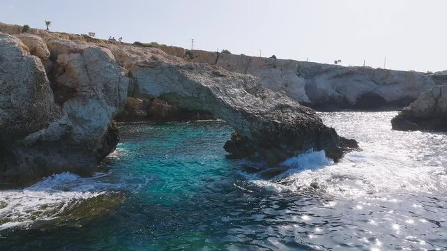 Aerial sweep of the Love Arch in Ayia Napa, Cyprus, with clear blue waters, gentle waves, and distant resorts along the coastline.
