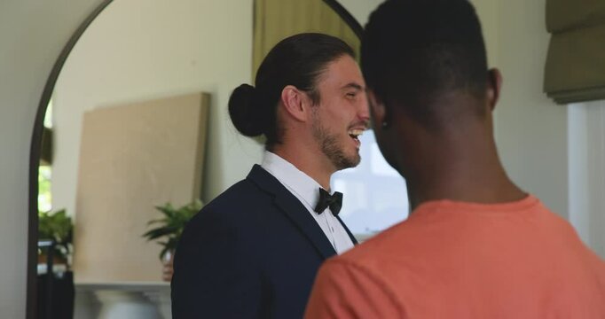 Diverse male friends entering fitting room hugging and welcoming tuxedoed man by arched mirror