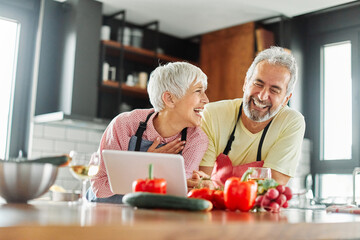 Portrait of happy senior mid aged mature couple prepering meal with fresh vegatebles and following internet instructions for a recipe on a tablet computer  or looking at video  or website app in kitch