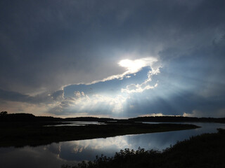 A bright sun breaks through the clouds, and emits an array of sunbeams upon the dark wetlands...