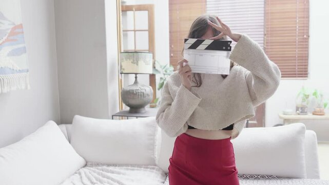 Woman holding a clapperboard in front of her face, playfully posing in a cozy living room. The scene is bright and airy, suggesting a relaxed and creative atmosphere.
