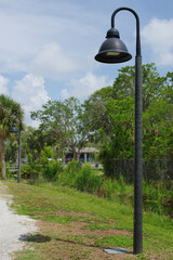 Street Lamp Along a Park Path with Trees and Grass in Daylight. Black  metal lamp stands on a serene path bordered by lush greenery and trees, with rural surroundings under a partly cloudy blue sky ev