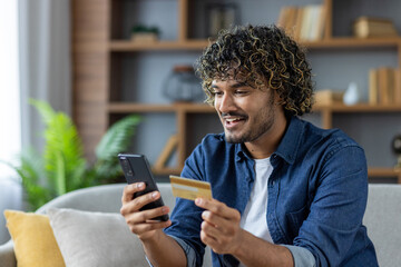 A smiling man is shopping online at home, using his phone and a credit card to make a purchase. The...